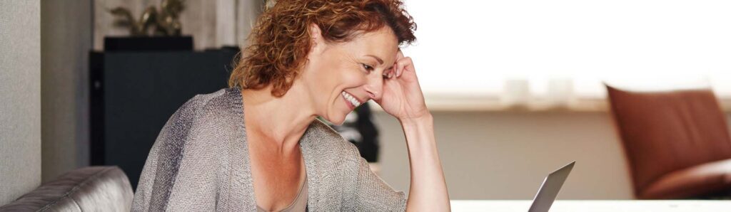 A woman smiles as she watches her laptop. This could represent the convenience of online therapy in Iowa. Learn more about online therapy for depression in Iowa and other services today!