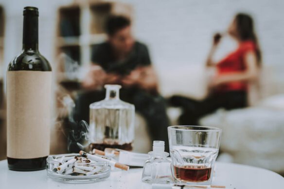 Wine and liquor next to an ash tray. A blurred couple talk in the back. Alcohol consumption and smoking can be signs of unhealed childhood wounds. A trauma EMDR therapist can help you and your relationships in West Des Moines, IA 50266